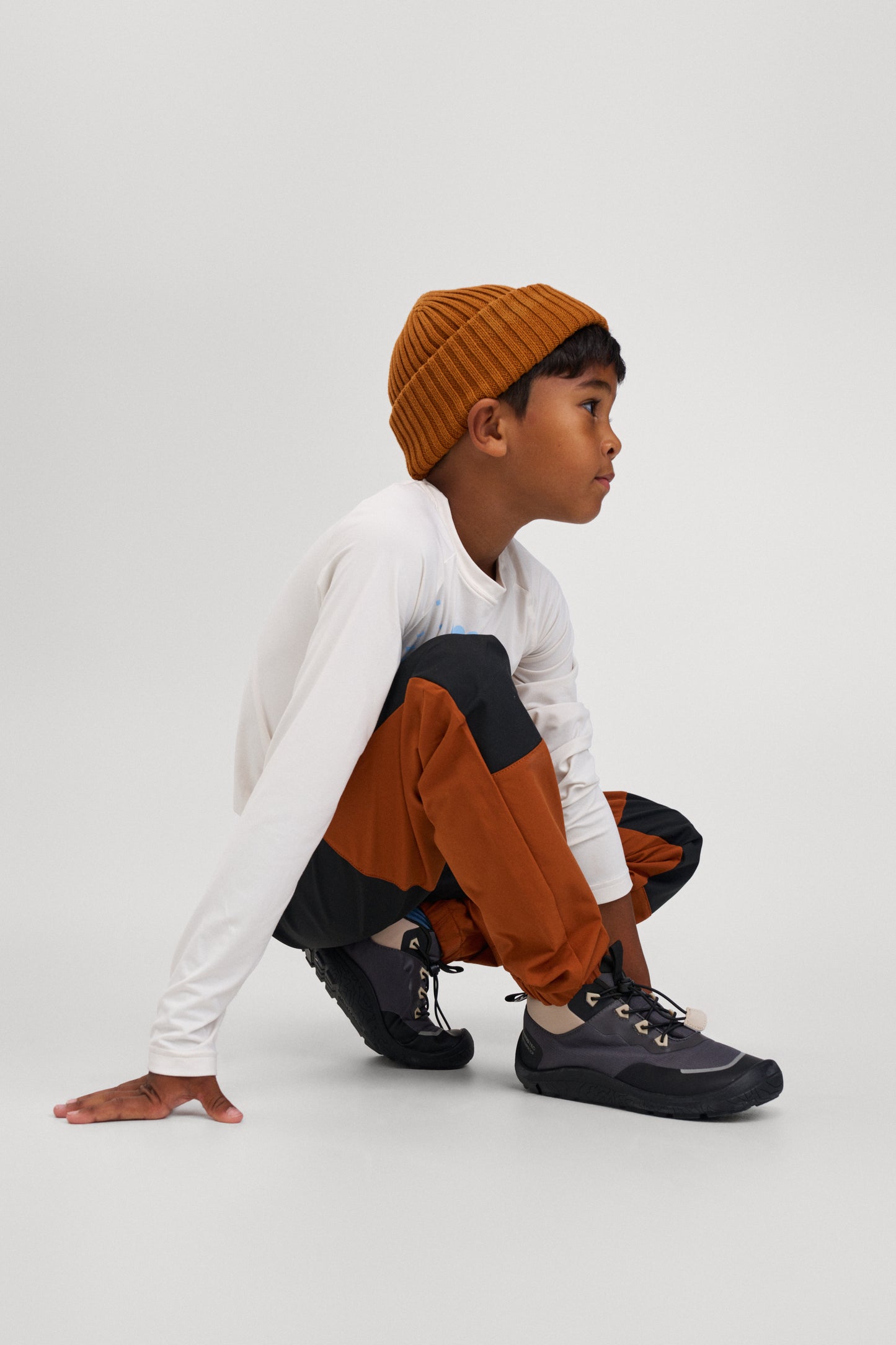 Child wearing a mustard beanie, white shirt, brown pants, and black shoes on a white background