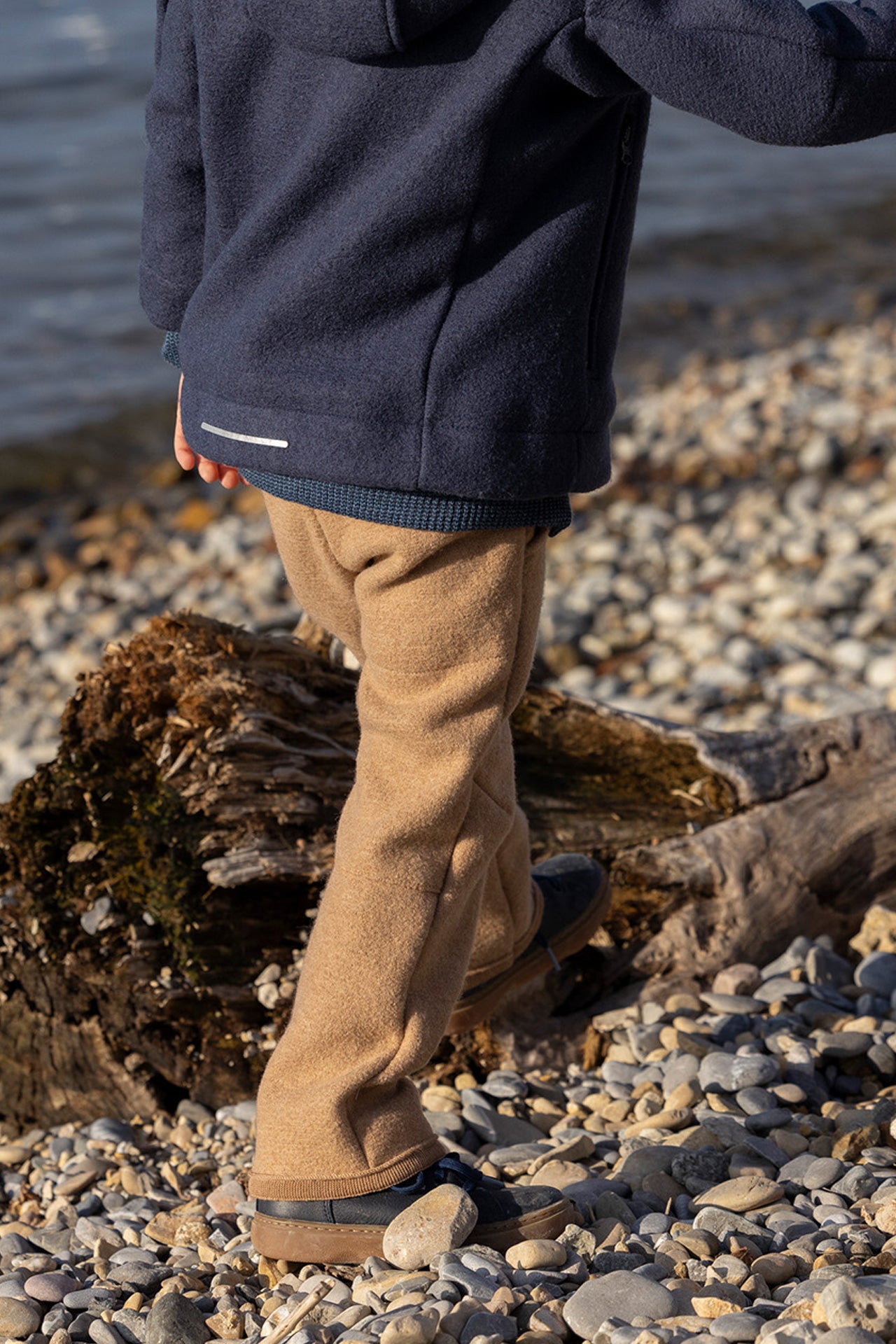 Person wearing a navy jacket and tan pants standing on a pebbly beach.