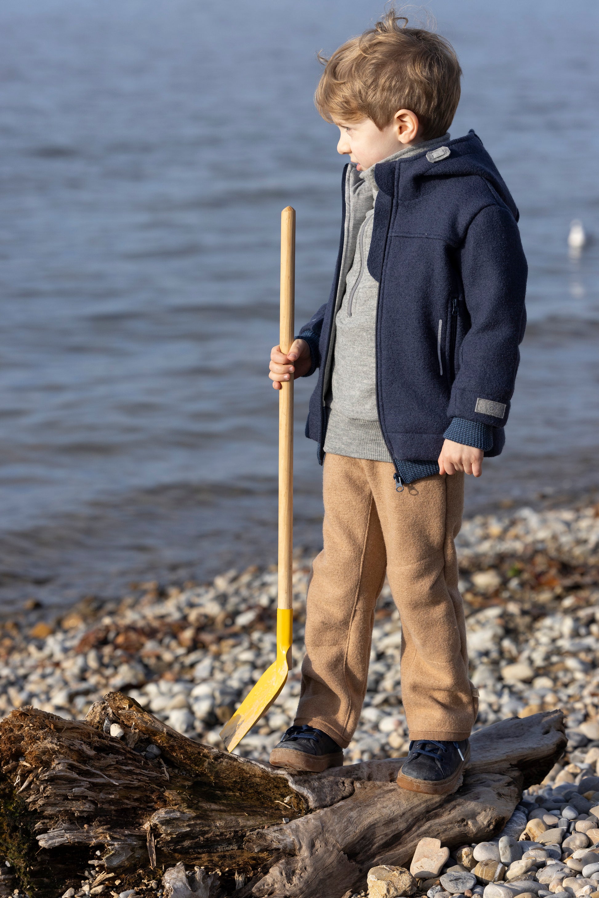 Child standing on a pebbly beach holding a yellow spade, wearing a navy coat and brown wool pants.